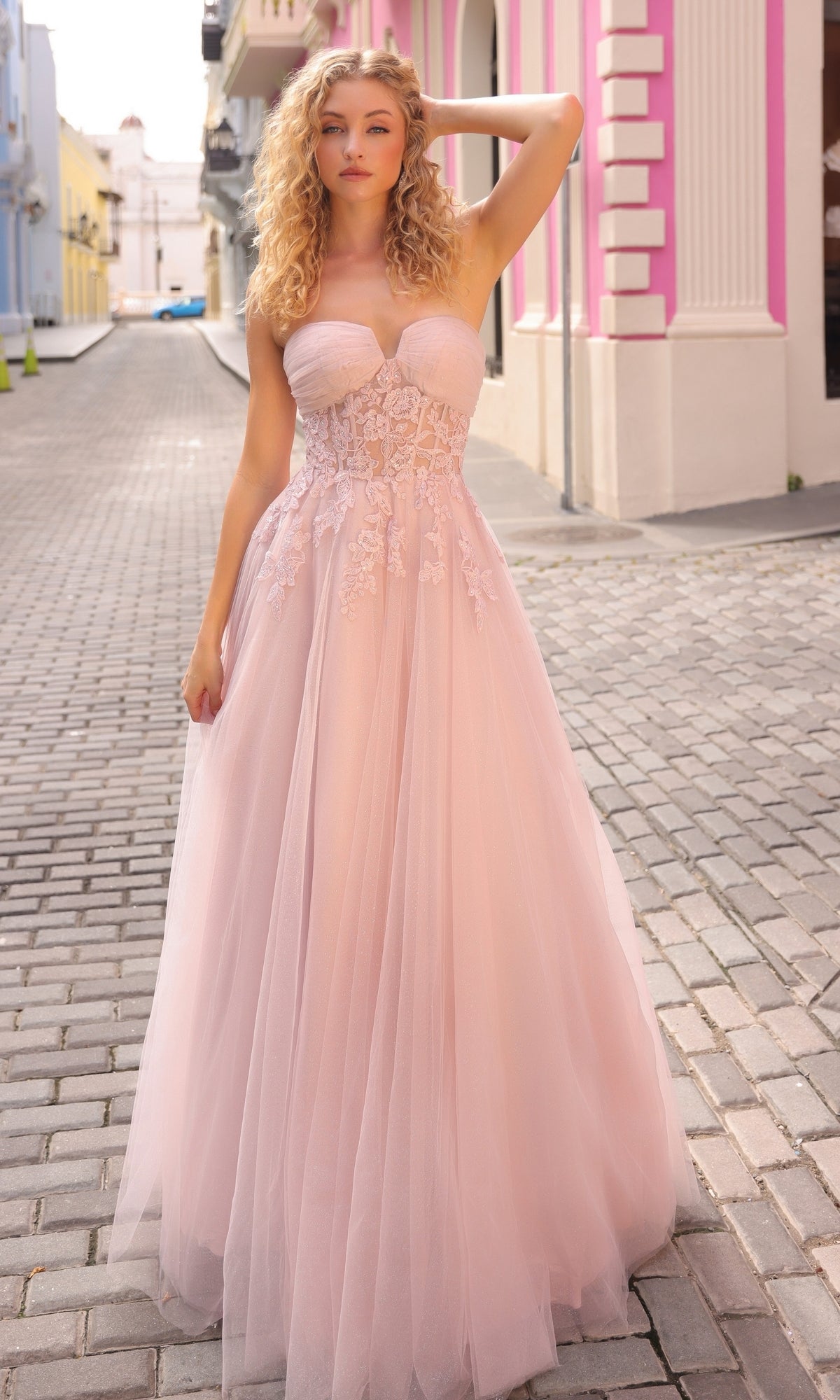 A woman stands on a cobblestone street wearing the Strapless Sheer-Waist Long Prom Ball Gown T1326, featuring light pink tulle and lace detailing on the bodice.